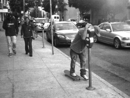 A grainy black and white photo of a sidewalk scene. In the photo a man has one foot on a skateboard and the other foot on the ground, he is hunched over and leaning on a parking meter for support as two friends watch him from behind. The man is learning how to skateboard and his friends are having fun watching him.