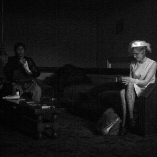 A grainy black and white photo of a woman in sitting on a couch. The lighting and couch are all very dark, but the woman is wearing a white dress and white hat, which makes her pop out from the rest of the scene.