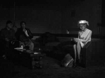 A grainy black and white photo of a woman in sitting on a couch. The lighting and couch are all very dark, but the woman is wearing a white dress and white hat, which makes her pop out from the rest of the scene.
